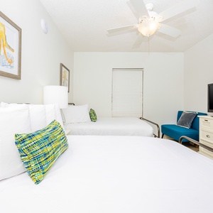Cheerful guest room featuring two queen beds, colorful decor, a TV, and a cozy seating area for relaxation