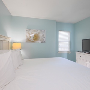 This bedroom features soft blue walls, a cozy white bed, and a TV, perfect for relaxing after a day at the beach