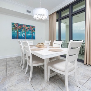 This dining area features a white table with six chairs, colorful palm art, and large windows offering a beachfront view