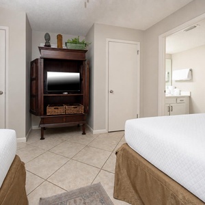 Bright guest bedroom featuring two double beds, wooden armoire with a TV, and a view into the bathroom area with fresh towels