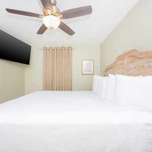 Bright guest bedroom featuring a king bed with a rustic wooden headboard, a ceiling fan, and a wall-mounted TV