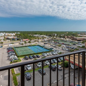 The balcony view showcases nearby tennis courts and a bustling parking area, framed by a vibrant blue sky and lush greenery