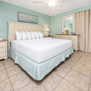 The bedroom features a king bed with plush pillows, a mounted TV, and soft teal walls, creating a serene retreat
