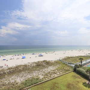 Expansive beach view showcases white sand and vibrant umbrellas, with a glimpse of the pool area framed by lush greenery