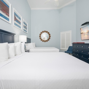 Bright and airy guest bedroom featuring two queen beds, a mounted TV, and coastal artwork for a relaxing atmosphere