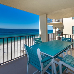 A bright balcony features a teal dining set with a stunning view of the white sand beach and turquoise waters below