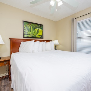 This bedroom features a large bed with crisp white linens, a tropical wall art piece, and wooden accents for a relaxing stay