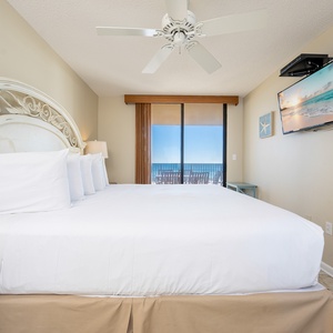 Bright master bedroom with a king bed, a mounted TV, and sliding glass doors leading to a private balcony with beach views