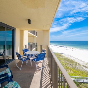 A spacious balcony features a round dining table and blue chairs, offering a serene view of the beach and turquoise waters