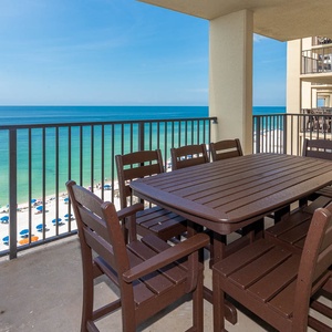 A spacious balcony features a wooden dining set with a view of the beach, showcasing vibrant umbrellas and clear blue waters