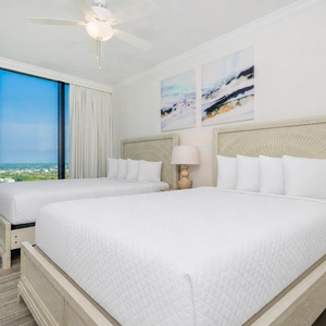 Rest in this guest bedroom featuring two cozy queen beds, a stylish lamp, and large windows showcasing beach views