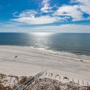 A serene beachfront view showcases soft white sand and gentle waves under a bright blue sky with scattered clouds