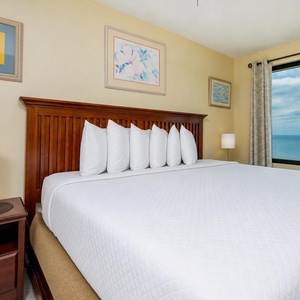 This bedroom features a king-sized bed, soft white linens, and a stunning beachfront view through large windows