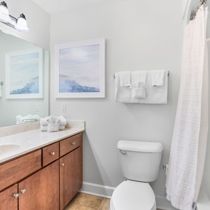 This bathroom features a stylish vanity with warm wood cabinetry, tub-shower, fresh white towels, and calming blue artwork