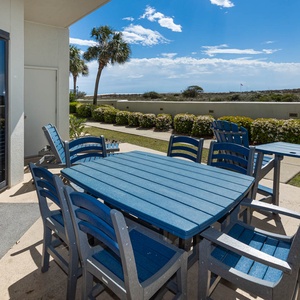 Outdoor patio area features a large blue table surrounded by matching chairs, with views of lush greenery and the beach