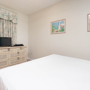 This cozy bedroom features a TV on a wicker dresser, soft pastel curtains, and charming artwork for a relaxing stay