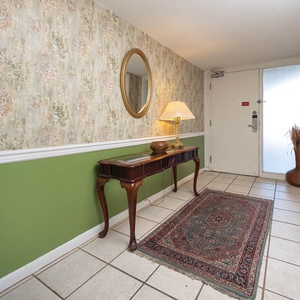 The entryway features floral wallpaper, a decorative mirror, a wooden table with a lamp, and a welcoming area rug