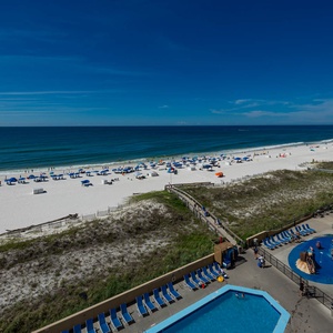 The view showcases a vibrant beach with white sand, lined with colorful umbrellas and a pool area below, inviting relaxation
