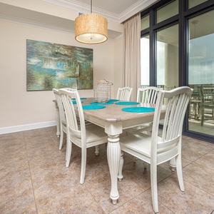 This dining area features a wood table with turquoise accents and six white chairs, and offers a view through large windows