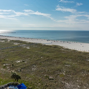 Expansive view of the beach with white sand and colorful umbrellas, framed by lush greenery and a sparkling pool area