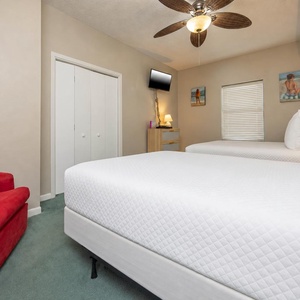 Bright guest bedroom featuring two double beds, a cozy red chair, and beach-themed artwork, creating a relaxed atmosphere