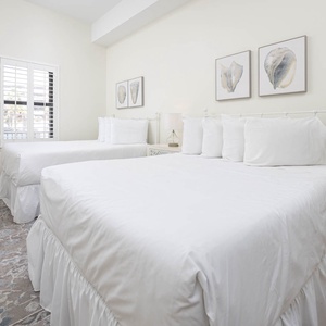 Two double beds with plush white bedding, complemented by coastal artwork and a cozy area rug in this guest bedroom
