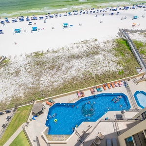 Aerial balcony view showcasing the vibrant pool area with loungers, leading to the white sand and colorful umbrellas