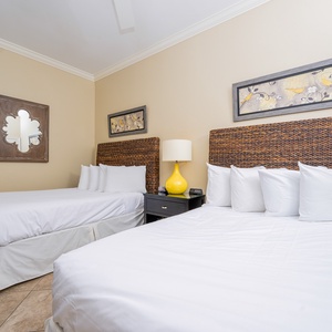 This guest bedroom features two queen beds with woven headboards, a bright yellow lamp, and charming artwork on the walls