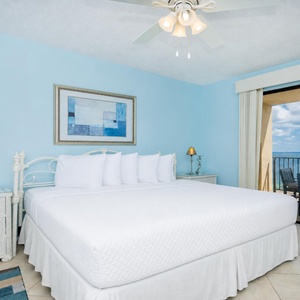 Brightly decorated master bedroom features a king bed, coastal art, and access to a balcony with a breathtaking beach view