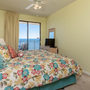 This master bedroom features a king bed, a TV, and a balcony door showcasing the beachfront view