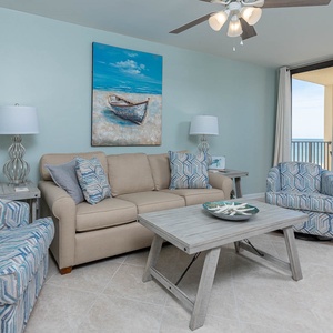The living area features a beige sofa with queen sleeper, patterned chairs, coastal art, and a beach front view