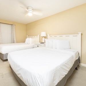 Bright and airy guest room featuring two queen beds, a ceiling fan, and a bedside table with a lamp for a restful stay