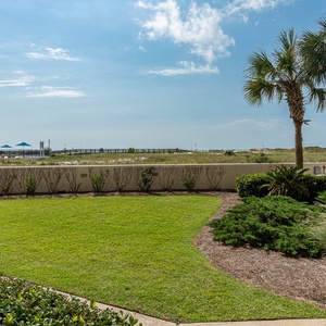 Lush green grass and palm trees frame a tranquil view of the beach, with a gazebo and lounge area visible in the distance