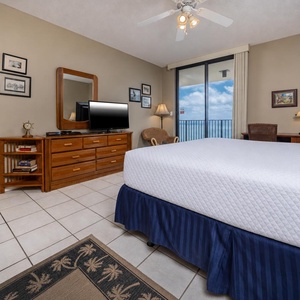 Comfortable master suite features a king bed, dresser with TV, and a chair, with a balcony offering a view of the beach