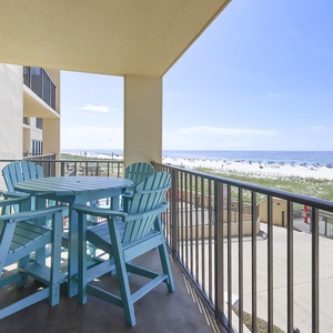 The balcony features a round table with bright turquoise chairs, offering a view of the beach and colorful umbrellas below