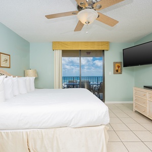 Cozy master bedroom featuring a large king bed, a wall-mounted TV, and sliding doors leading to a balcony with beach views