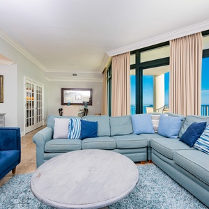 The living area features a plush sectional sofa, a round coffee table, and stunning beach views through large windows