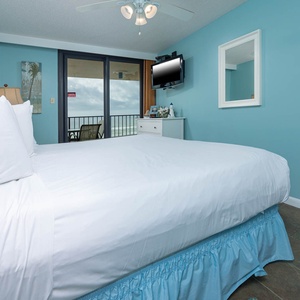A bright master bedroom features a king bed with a blue skirt, a TV on the wall, and sliding doors leading to the balcony