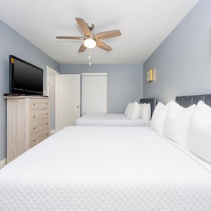 A guest room features two neatly made full beds with plush white pillows, a flat-screen TV, and soft blue walls