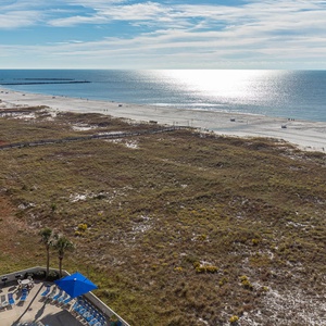 Stunning view of the beach with glistening water, framed by lush greenery and a pool area with blue umbrellas and loungers