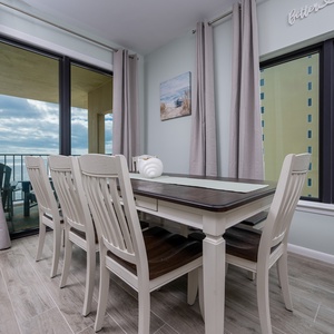 This dining area features a stylish table with white chairs, large windows offering a glimpse of the beautiful beach view