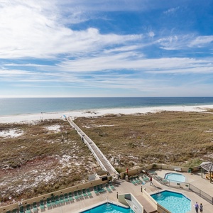 Stunning view of the beach with a wooden walkway leading to the shore, framed by a pool area and lush greenery