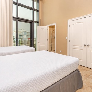 This bright bedroom features two full beds, a colorful dresser, and a TV, with large windows offering a view of the outdoors
