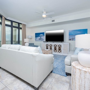 This living area features a large TV, light-colored furniture, and stunning beachfront views through expansive glass windows