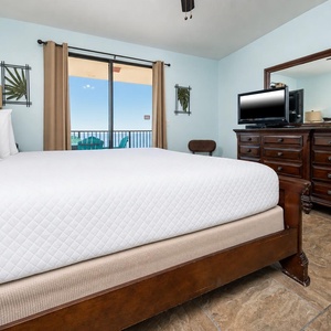 The inviting master bedroom features a king bed, a wooden dresser with a TV, and a balcony view showcasing outdoor seating