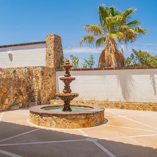 A sunny courtyard features a stone fountain with elegant tiers, palm trees, and colorful stonework creating a tranquil outdoor space.