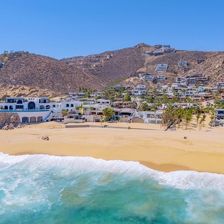 Beachfront hillside neighborhood with residential properties overlooking sandy shore and turquoise ocean waters, offering a scenic coastal location.