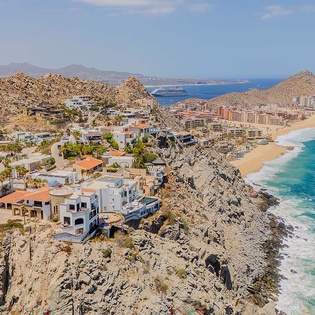 Aerial view of a stunning coastal neighborhood with dramatic rocky cliffs, beachfront properties, and pristine turquoise waters framed by golden sand and distant mountains.