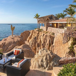 Oceanfront villa perched on dramatic cliffs, with palm trees swaying above. Stone terraces and outdoor lounges frame unobstructed views of the turquoise sea and golden coastline.