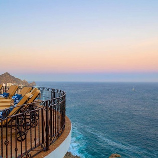 Breathtaking oceanfront balcony showcasing a stunning sea view at sunset, with poolside loungers poised above turquoise waters and distant sailboats dotting the horizon.